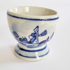 May include: A small, white ceramic egg cup with a blue windmill and landscape design. The cup has a pedestal base and a blue rim. The design is reminiscent of Delftware pottery.