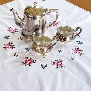 Könnte beinhalten: Ein silbernes Teeservice mit Teekanne, Sahnekännchen und Zuckerdose steht auf einer weißen Tischdecke. Die Tischdecke ist mit rosa, blauen und gelben Blumenmustern bestickt. Das Set steht auf einem Holztisch.