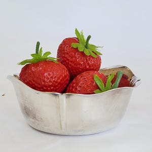 May include: A small, polished silver-toned bowl with a scalloped rim containing three bright red artificial strawberries with green plastic stems and leaves. The bowl has a smooth, reflective metallic surface and a rounded, shallow basin shape.