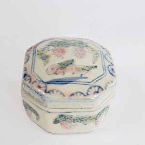 Könnte beinhalten: Eine kleine, weiße Porzellanbox mit Deckel. Die Box ist achteckig und hat ein florales Design in Rosa-, Blau- und Grüntönen. Der Deckel hat einen gewellten Rand.
