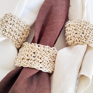 Peut inclure: Trois serviettes en tissu blanc, marron et crème sont maintenues par des ronds de serviette beiges crochetés. Les serviettes sont pliées et disposées pour mettre en valeur les anneaux. Les anneaux ont un aspect texturé et tissé. L'image est bien éclairée.