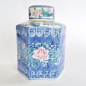 Pode incluir: Um pote de cerâmica hexagonal com tampa removível, apresentando um fundo azul decorado com motivos florais cor-de-rosa e folhagem verde. O recipiente exibe um padrão repetitivo de flores estilizadas e desenhos de bordas complexos em branco e azul.