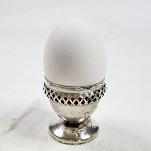 May include: A white egg in a silver egg cup. The egg cup has a decorative, openwork design around the top and a small, round base. The egg is smooth and oval-shaped, and the cup appears to be made of silver-toned metal.