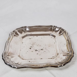 Può includere: Un vassoio rettangolare color argento con un bordo decorativo smerlato. Il vassoio ha una superficie leggermente appannata, che mostra segni di età. Il design è semplice ma elegante, adatto per servire.