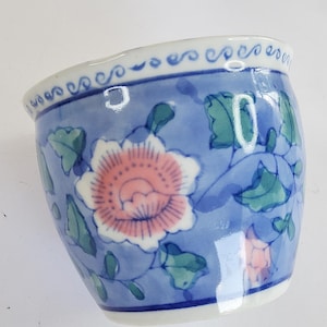 Könnte beinhalten: Kleine blaue Keramikschale mit floralem Design. Die Schale zeigt eine rosa-weiße Blume mit grünen Blättern auf blauem Hintergrund. Der Rand hat einen weißen Rand mit sich wiederholendem Muster. Ein dekoratives Stück.