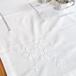以下が含まれることがあります： 白い刺繍入りのリネンテーブルクロス。花柄と幾何学模様が施されています。取っ手付きの銀色のカップが3つテーブルクロスの上に置かれています。テーブルクロスは木製の表面にあります。
