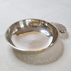 Könnte beinhalten: Eine kleine, runde, silberfarbene Schale mit einem muschelförmigen Griff. Die Schale hat eine reflektierende Oberfläche und steht auf einer weißen, strukturierten Oberfläche. Die Schale dient wahrscheinlich zum Servieren oder Ausstellen.