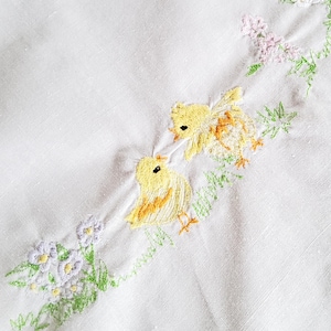 Könnte beinhalten: Weißer Stoff mit gestickten gelben Küken und floralen Akzenten in Grün, Lila und Gelb. Die Stickerei zeigt zwei Küken mit detaillierten Federn und Schnäbeln, umgeben von zarten Blumen und Blättern, die ein Frühlings- oder Osterthema schaffen.