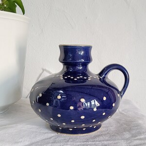 Może przedstawiać: Ciemnoniebieski dzbanek ceramiczny z zaokrąglonym korpusem i pojedynczym uchwytem. Powierzchnia ozdobiona jest małymi, równomiernie rozmieszczonymi białymi kropkami. Dzbanek posiada krótką, cylindryczną szyjkę oraz gładkie, błyszczące wykończenie.