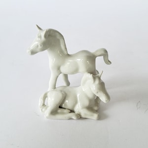 Könnte beinhalten: Zwei kleine, cremefarbene Keramik-Einhornfiguren. Ein Einhorn steht aufrecht, das andere liegt. Beide haben ein einzelnes Horn und sind vor einem weißen Hintergrund platziert. Die Figuren sind wahrscheinlich Dekorationsartikel.