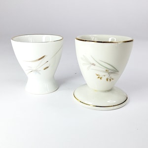 Puede incluir: Dos hueveras de cerámica blanca con ribete dorado. Una taza tiene un diseño simple de líneas doradas y grises, mientras que la otra presenta un diseño floral en oro, gris y verde. Las hueveras están sobre un fondo blanco.