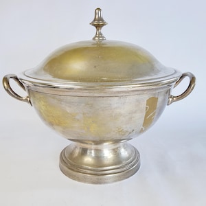 Könnte beinhalten: Eine silberne Servierschüssel mit Deckel und zwei Griffen. Die Schüssel steht auf einem Sockel.
