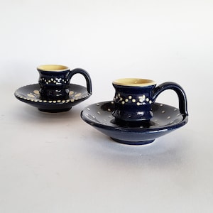 Può includere: Due portacandele in ceramica blu scuro con manici, ciascuno caratterizzato da una base ampia e poco profonda e una parte superiore cilindrica. Le superfici esterne sono decorate con piccoli punti giallo chiaro in bande.