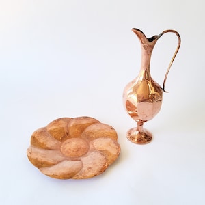 May include: A polished copper pitcher with a slender neck, curved handle, and faceted body, positioned next to a circular wooden tray featuring a carved, multi-petaled flower design with a textured, matte finish and warm brown tones.