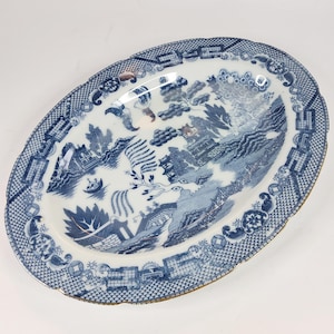 Może przedstawiać: Owalny półmisek ceramiczny z niebiesko-białym wzorem willow. Projekt przedstawia krajobraz z mostem, pagodą, drzewami i małą łodzią na wodzie. Brzeg zdobi geometryczny wzór kratki z kwiatowymi akcentami.