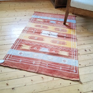 May include: A rectangular area rug featuring a horizontal striped pattern in shades of terracotta, mustard yellow, and light blue. The rug is decorated with white geometric motifs, including diamonds and zigzag lines, and has a thin white border.