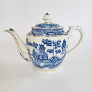Könnte beinhalten: Eine weiße Porzellan-Teekanne mit blau-weißem Willow-Muster. Die Teekanne hat einen gebogenen Ausguss, einen abgerundeten Körper und einen Deckel mit Knauf. Goldbesatz akzentuiert den Ausguss und den Sockel. Der Griff ist eine gebogene Schlaufe.