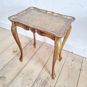 May include: An antique gold-painted side table with a rectangular top and four curved legs. The table's top features a decorative floral design in shades of pink and brown. The table is approximately 24 inches tall.