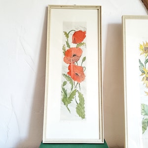 Könnte beinhalten: Ein vertikaler, rechteckiger gerahmter Druck mit einer botanischen Illustration von drei leuchtend roten Mohnblumen mit grünen Blättern und Stängeln. Das Kunstwerk ist auf hellem Grund und in einem dünnen, hellen Holzrahmen gefasst.