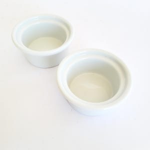 Op de afbeelding: Twee kleine, ronde, witte keramische ramequins met een gladde, glanzende afwerking en brede, uitlopende randen. Deze keukenvaten hebben diepe, cilindrische interieurs en zijn ontworpen voor individuele porties voedsel.