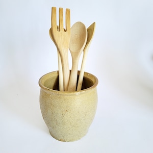 Può includere: Un portautensili in ceramica color beige chiaro maculato contenente tre utensili da cucina in legno chiaro, tra cui una forchetta a tre rebbi, un cucchiaio arrotondato e una spatola appuntita, disposti verticalmente nel contenitore.