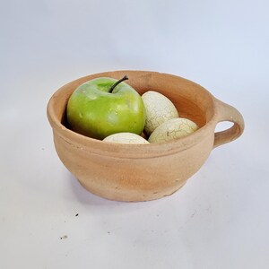 Puede incluir: Un cuenco rústico de color terracota con asa, que contiene una manzana verde y varios huevos decorativos moteados blanquecinos. El cuenco tiene un acabado sencillo, sin esmaltar, lo que sugiere una calidad artesanal. La disposición evoca la simplicidad natural.