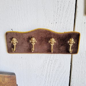 Könnte beinhalten: Ein Vintage-Wandgarderobe mit vier Messinghaken. Das Gestell hat eine braune Samtrückseite mit goldenem Besatz. Jeder Haken ist mit einem dekorativen Gesicht verziert. Die Garderobe ist auf einer weißen Holzoberfläche montiert.