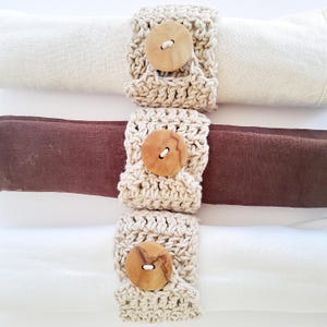 Peut inclure: Trois serviettes enroulées, crème, bordeaux et blanche, chacune maintenue par un anneau de serviette crocheté. Les ronds de serviette sont beiges et comportent un grand bouton en bois rond.