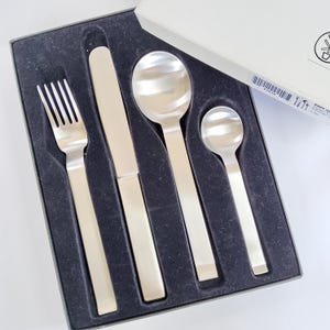 May include: A stainless steel flatware set, including a fork, knife, and two spoons, presented in a black velvet-lined box. The silverware has a modern design with rectangular handles and a polished finish. The box is partially open, revealing the contents.