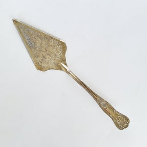 May include: A gold-toned metal cake server featuring an intricate engraved floral pattern on the triangular blade. The handle has a decorative embossed shell motif at the end and a slender, grooved shaft connecting to the wide, serrated-edge blade.