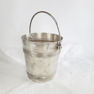 Könnte beinhalten: Ein silberfarbener Metalleimer mit einem glatten, konischen Körper und horizontalen Rillendetails an den oberen und unteren Rändern. Der Eimer verfügt über einen dünnen, gebogenen Metallgriff, der mit runden Nieten an den Seiten befestigt ist.
