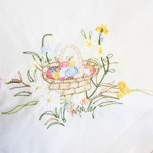 Könnte beinhalten: Gestickter Osterkorb gefüllt mit bunten Eiern, umgeben von Gänseblümchen, Narzissen und Grünzeug. Der Korb ist mit einem Henkel gewebt, und die Stickerei ist in verschiedenen Farben auf weißem Grund ausgeführt.