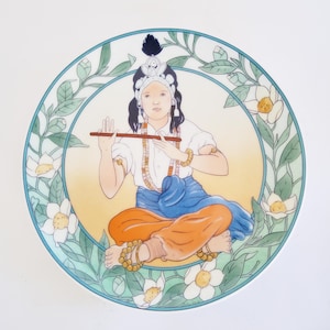 May include: A decorative plate depicting a seated figure playing a flute. The figure wears a crown, jewellery, and traditional clothing in white, blue, and orange. The plate is framed by a floral design with white flowers and green leaves.