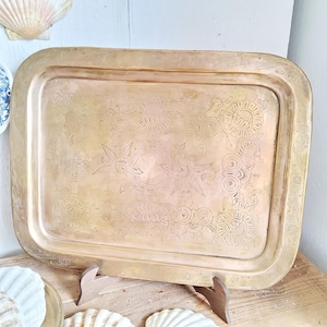 May include: A rectangular brass tray with rounded corners, featuring an engraved floral and sunburst design. The tray is supported by a wooden stand and is displayed on a wooden surface. Decorative seashells are visible in the background.