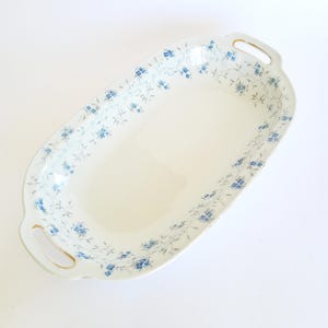 May include: Oval serving platter with a white base and delicate blue floral pattern around the rim. The platter has gold-trimmed handles on each end, offering a touch of elegance. A classic piece for serving appetizers or desserts.