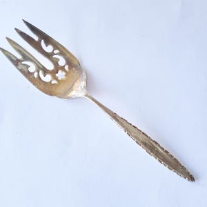 May include: Silver serving fork with a decorative openwork design on the tines. The fork has four tines and a long, slender handle with a textured edge. The utensil is likely vintage or antique.