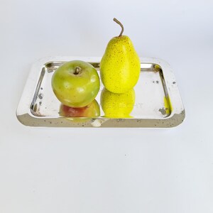 May include: A silver rectangular tray with rounded corners holds a green apple and a yellow pear. The apple is on the left, and the pear is on the right. The tray has some wear and tear. The word "FRUECHT" is visible on the tray.