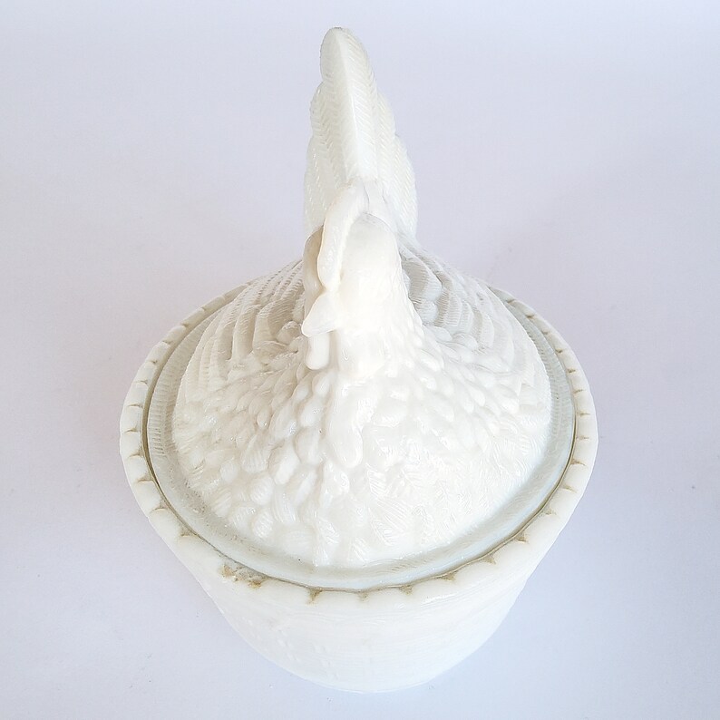 May include: White ceramic chicken-shaped butter dish with a detailed design. The dish has a lid shaped like a chicken's head and tail feathers.