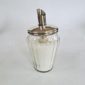 Peut inclure: Un sucrier en verre transparent avec un couvercle et un bec verseur argentés. Le pot en verre présente des rainures verticales et est rempli de sucre granulé blanc. Le bec verseur est incliné et le couvercle est circulaire.