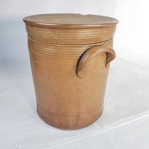 Könnte beinhalten: Ein zylindrischer Steinguttopf in einem beigefarbenen Farbton mit passendem Deckel. Er hat einen gebogenen Griff und horizontale geriffelte Details um den oberen und unteren Rand. Der Deckel sitzt bündig, und das Stück hat ein rustikales, gealtertes Aussehen.