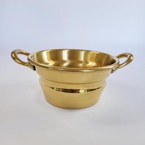 Op de afbeelding: Een gepolijste goudkleurige metalen kom met een glad, reflecterend oppervlak en een brede opening. De kom is voorzien van twee gebogen handgrepen aan weerszijden, elk met een geribbelde textuur, en een afgeronde basis die naar onderen toe afloopt.
