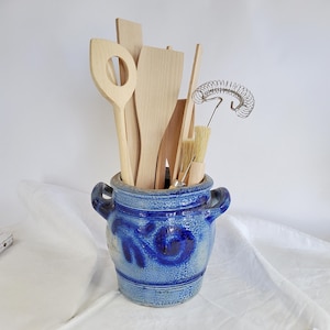 May include: A blue glazed ceramic crock with two side handles containing a collection of light-colored wooden kitchen utensils, including a slotted spoon, a flat spatula, and a metal wire whisk with a wooden handle standing upright.