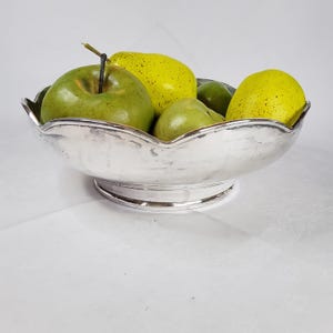 Puede incluir: Un cuenco plateado con borde festoneado lleno de manzanas verdes y peras amarillas. El cuenco tiene una base de pedestal y un acabado pulido, creando una superficie reflectante. La fruta está dispuesta para llenar el cuenco, creando una naturaleza muerta.