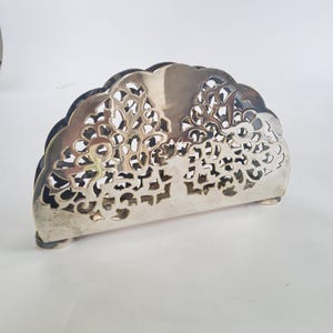May include: A silver-colored napkin holder with a decorative, openwork design. The holder has a semi-circular shape with a scalloped edge and rests on small feet. The intricate pattern adds an elegant touch to any table setting.