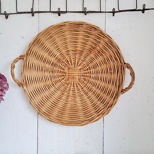 May include: A round, light brown woven wicker tray featuring a flat, circular base with a tight, concentric spiral pattern and two sturdy, braided handles attached to opposite sides. The tray is constructed from natural, flexible plant fibers.