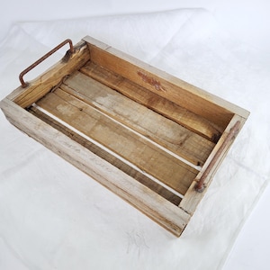 Könnte beinhalten: Eine rechteckige Holzkiste mit Metallgriffen. Die Kiste besteht aus hellen, verwitterten Holzbrettern. Die Metallgriffe sind rostbraun. Die Kiste hat ein rustikales, Vintage-Aussehen.