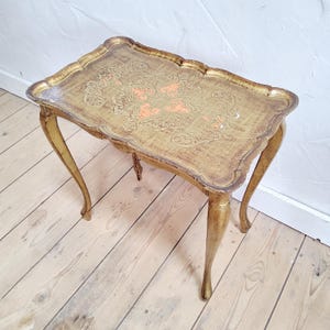 Puede incluir: Una mesa auxiliar rectangular con acabado en tono dorado y patas curvas. El tablero presenta un borde festoneado y un intrincado patrón en relieve en tonos dorados y naranja apagado, mostrando un aspecto envejecido en toda la superficie.