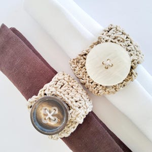 Peut inclure: Deux serviettes en tissu, une blanche et une marron, sont maintenues par des ronds de serviette en crochet. Chaque anneau comporte un gros bouton. Les anneaux en crochet sont beiges et les boutons sont blanc cassé et marron.