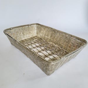 May include: A rectangular silver-colored metal basket with a woven design. The basket has a grid pattern on the bottom and a sturdy, rounded rim. The basket is empty and sits on a white surface.