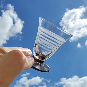 Peut inclure: Un verre transparent avec un dessus évasé et une base sombre, tenu contre un ciel bleu nuageux. Le verre présente des rayures blanches horizontales, créant un motif décoratif. Verrerie pour boissons.
